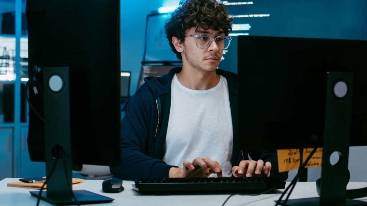 Male Software Engineer, Computer Programmer sits at desk Writing Code on Desktop computer with Dual Screens at Tech Office at Night. Professional Male IT expert working on Software Code and Program.