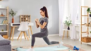 Active sporty young woman having fitness workout at home, doing front lunges on yoga mat in living room. Athletic smiling slim girl in sportswear strengthening her body, maintaining healthy routine.