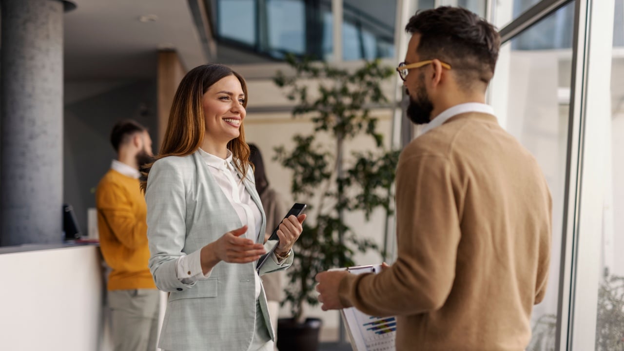 A professional businesswoman and a businessman engage in a friendly discussion in a modern, well-lit office space with large windows and greenery.