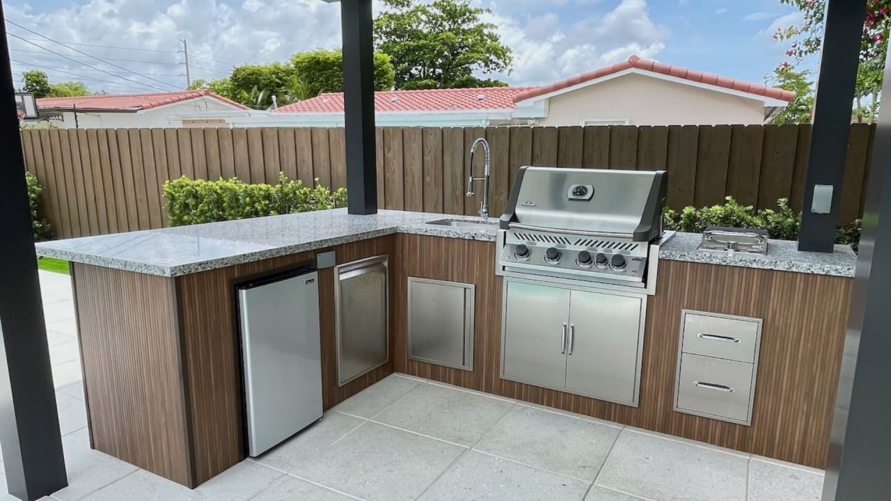 Modern outdoor kitchen with granite countertop, stainless-steel grill, mini fridge and sink under a pergola in a residential patio, ideal for alfresco cooking and entertaining.