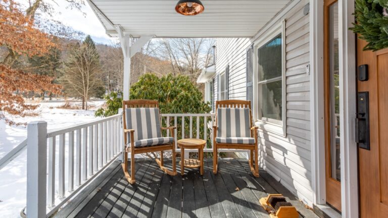 Roscoe, New York, USA – January 13, 2025: Front porch with wooden rocking chairs, striped cushions, small table, and snowy yard views at a home in a rural winter setting.