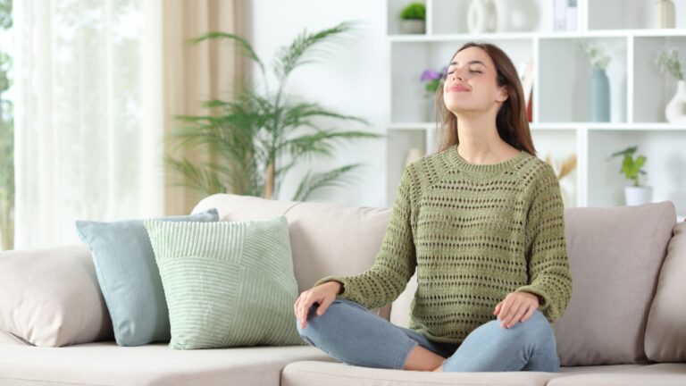 Happy woman in green breathing fresh air meditating sitting on a couch at home