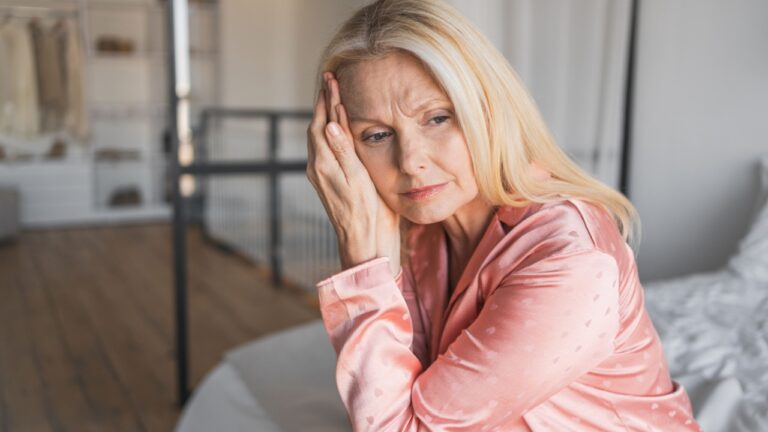 Lonely unhappy middle-aged woman feels depression sitting on bed. Unwashed female wearing pajama covering face with hand thinks about problems with work and finances suffering from headache