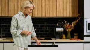 Caucasian middle aged woman standing in modern kitchen holding spoon and looking into open drawer, preparing for meal or organizing utensils, wooden cabinets in background
