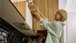 Middle aged Caucasian woman reaching for wicker basket on high kitchen shelf, standing sideways with short blond hair, preparing or organizing items on countertop in modern kitchen