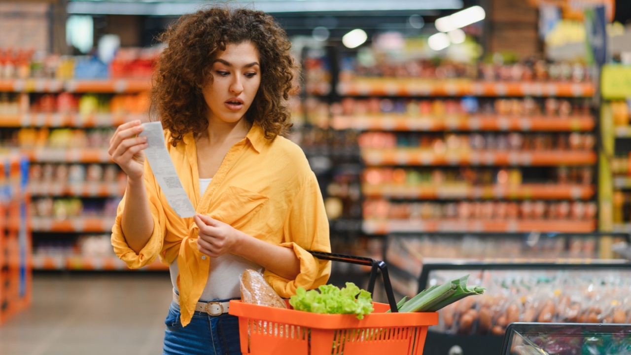 Woman holding a long shopping receipt, looking surprised and concerned about increasing food prices while shopping in a supermarket