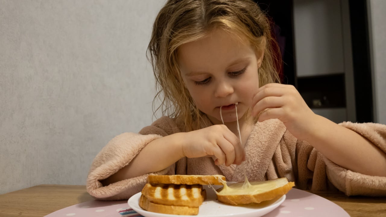 Cute little girl in bathrobe eating delicious grilled cheese sandwich with stretching cheese for breakfast at home