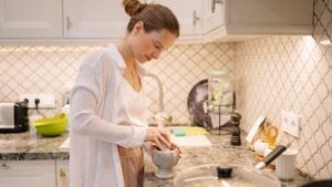 A Woman grinds spices in a mortar with a pestle in her kitchen. She wears light clothes. A pan sits on the stove. The kitchen is modern and well-lit.