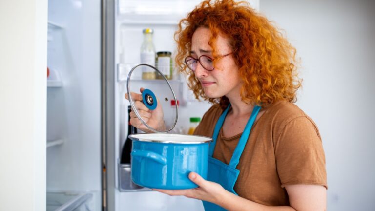 Disgusted redhead woman smelling spoiled food in a pot by the refrigerator. Young female reacting to bad smell of rotten leftovers in the kitchen.