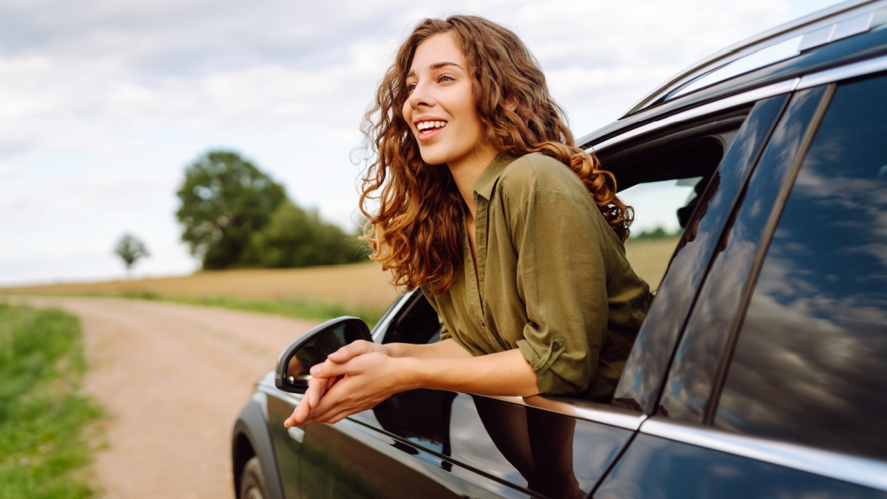 A beautiful woman leans out of a car window, enjoying a sunny day on a road trip. The young traveler feels the freedom of the outdoors. Concept of leisure and transportation.