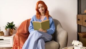 A young woman with red hair enjoys reading a book in her cozy winter attire at home.