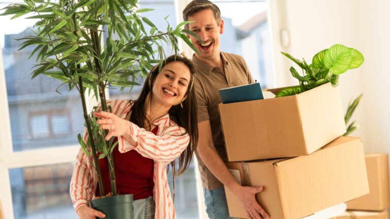 Two people are smiling while moving into a new home. One person holds a potted plant, while the other carries boxes. Sunlight comes through large windows in the background.