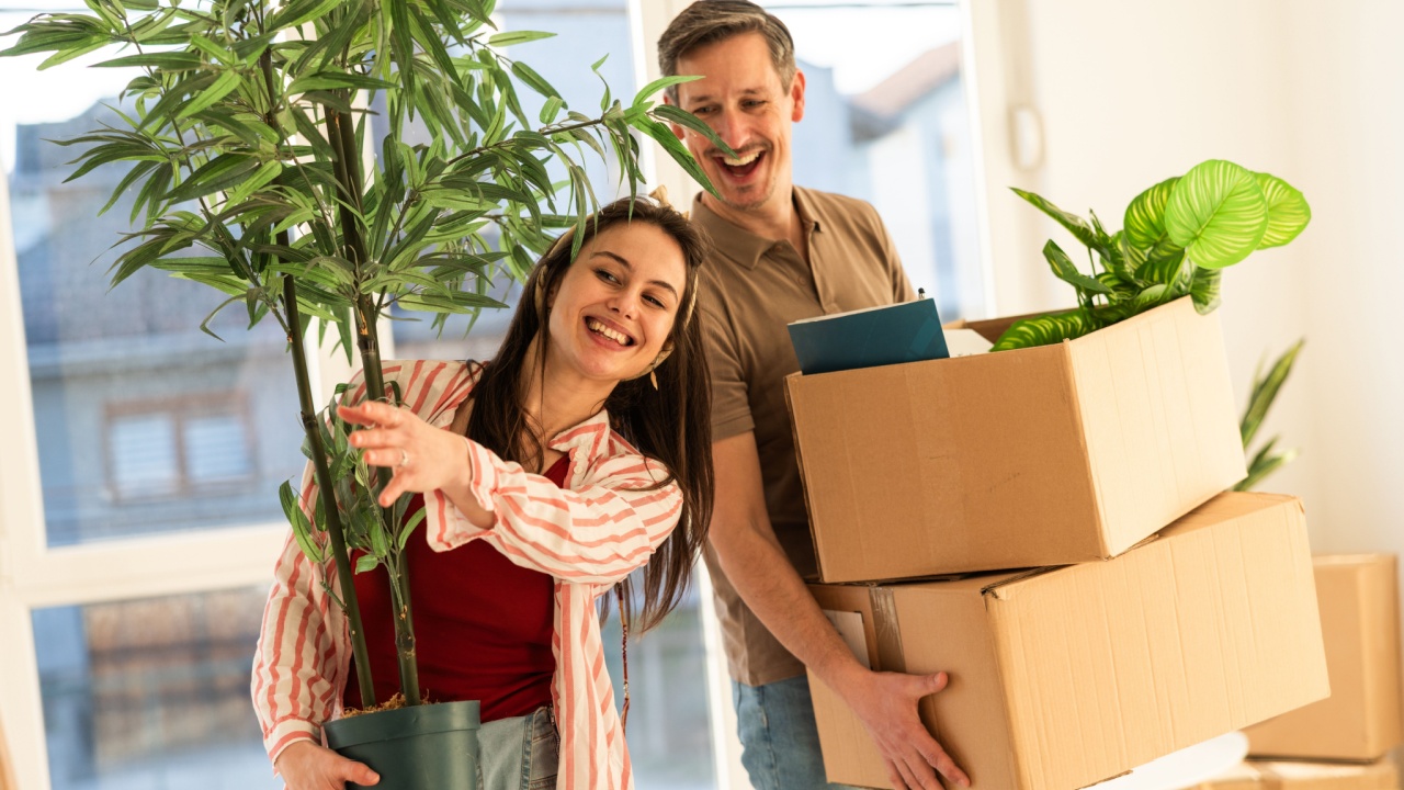 Two people are smiling while moving into a new home. One person holds a potted plant, while the other carries boxes. Sunlight comes through large windows in the background.
