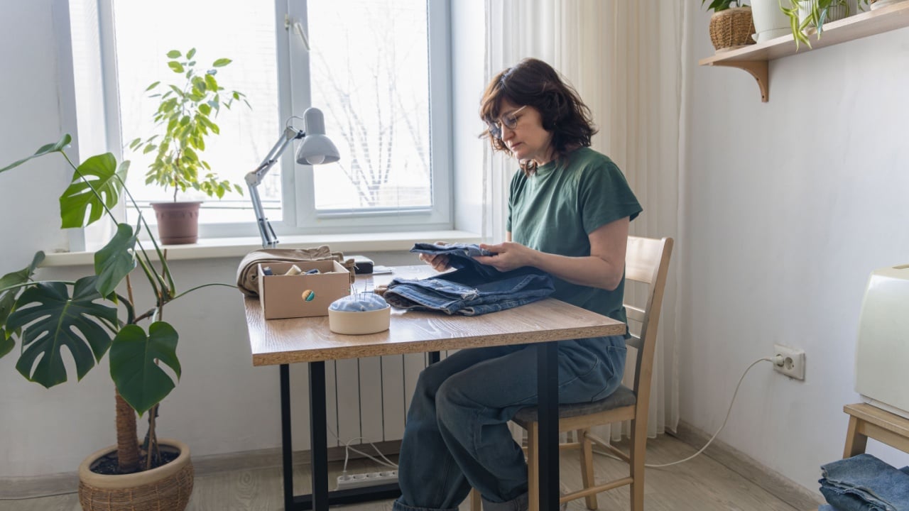 Woman sitting at a table near a window, holding denim fabric and preparing for a repair or upcycling sewing project. Woman examining denim fabric for sustainable fashion project