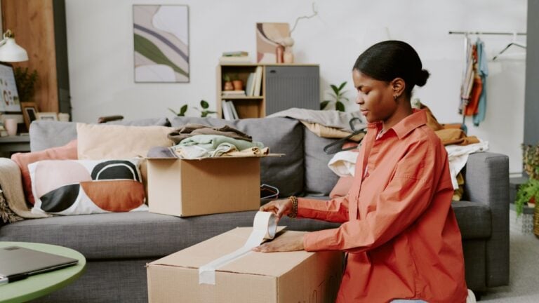 Black young adult woman sealing moving box while packing clothes at home, preparing relocation or online resale. Useful for moving services, decluttering, donation, ecommerce campaigns