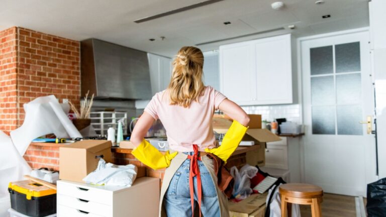 A woman with blonde hair, wearing yellow gloves and an apron, stands in a cluttered room filled with boxes and household items, facing away from the camera.