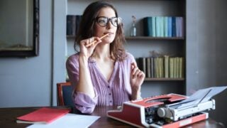 Portrait of a pensive mature authoress in glasses sitting at the table with papers and typewriter and looking away