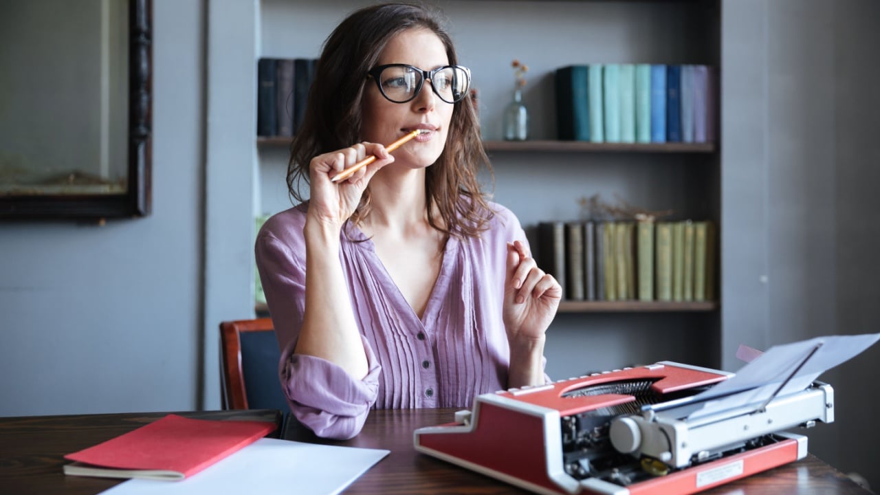 Portrait of a pensive mature authoress in glasses sitting at the table with papers and typewriter and looking away