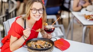Young woman in red dress eating sea Paella, traditional Valencian rice dish, sitting outdoors at the restaurant in Valencia, Spain