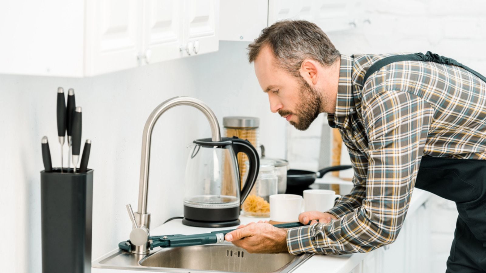 Side view of handsome plumber repairing tap with monkey wrench in kitchen