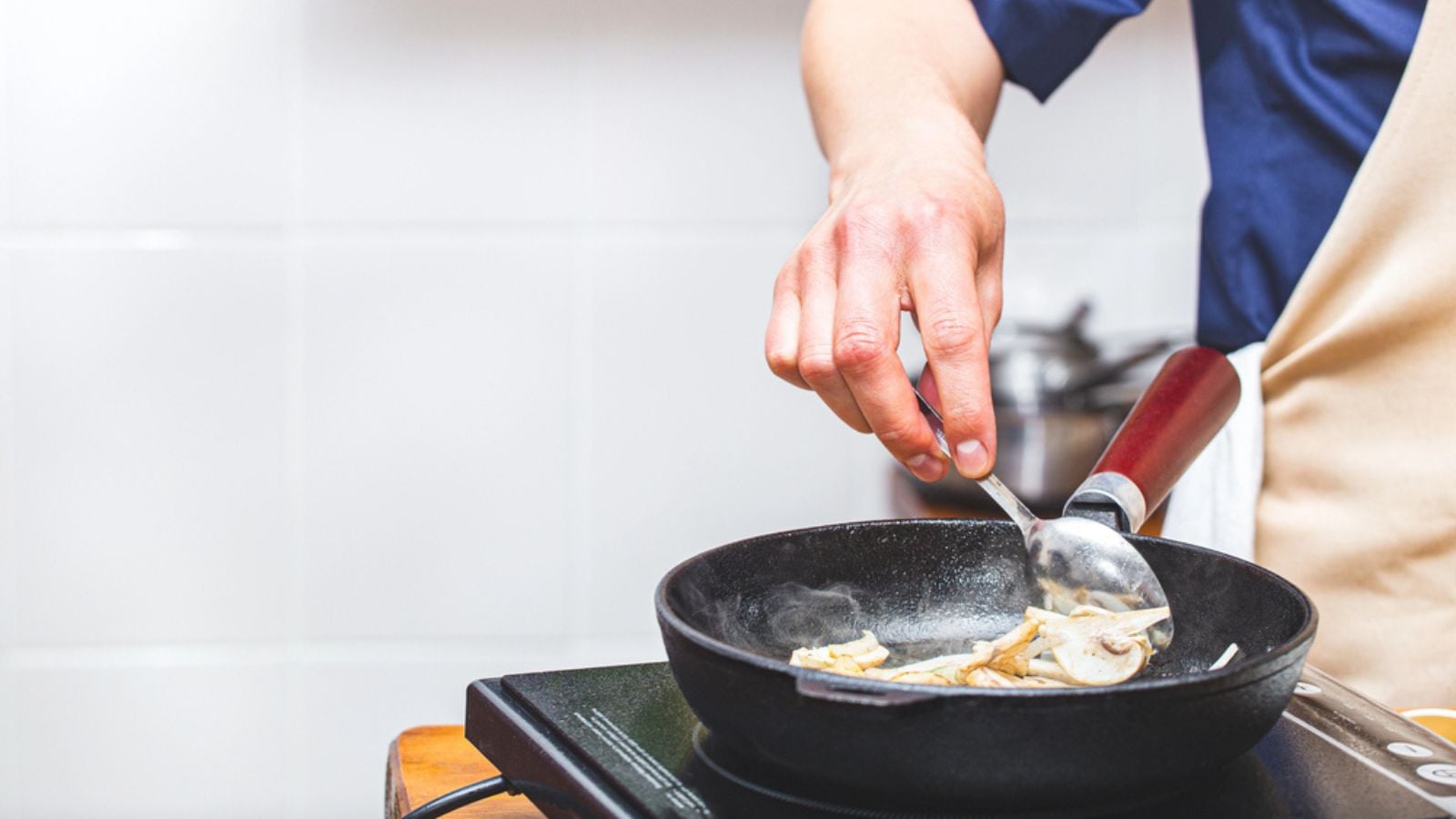 male chef in an apron fries mushrooms champignons in a black cast-iron pan.