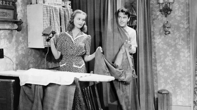 Man and woman standing in a kitchen while she is ironing his pants and he is behind a curtain