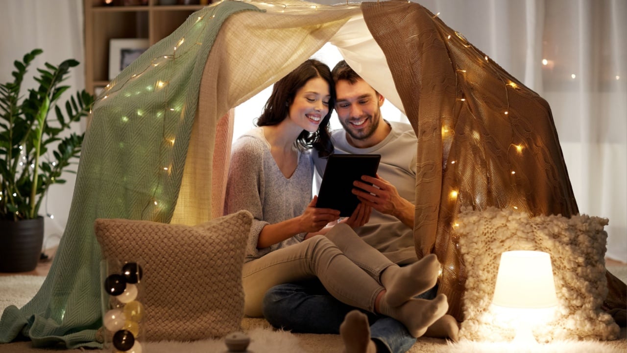 leisure, hygge, technology and people concept - happy couple with tablet pc computer in kids tent at home