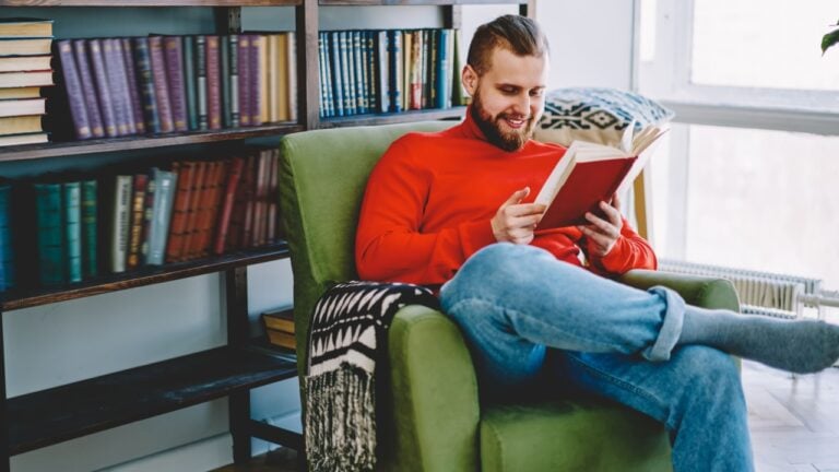 Positive caucasian male resting in comfortable armchair with interesting novel satisfied with free time,smiling hipster guy enjoy interesting book from home library collection in living room