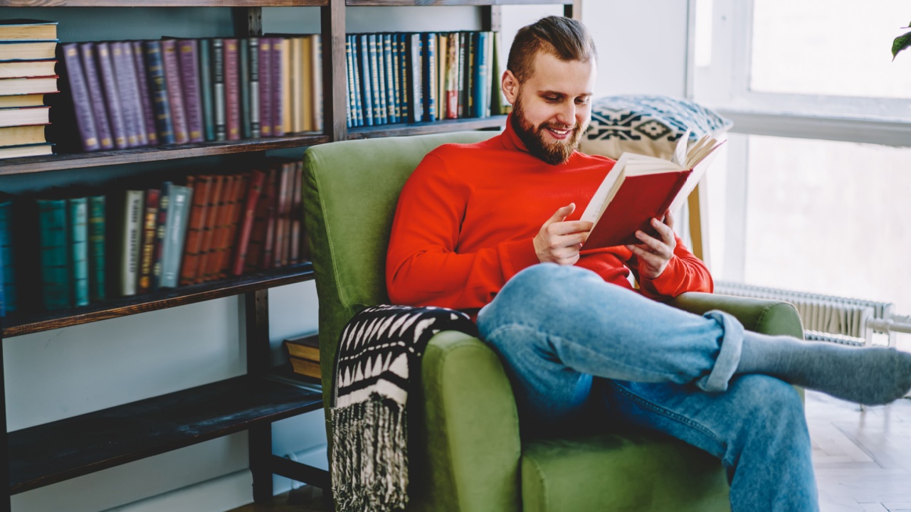 Positive caucasian male resting in comfortable armchair with interesting novel satisfied with free time,smiling hipster guy enjoy interesting book from home library collection in living room