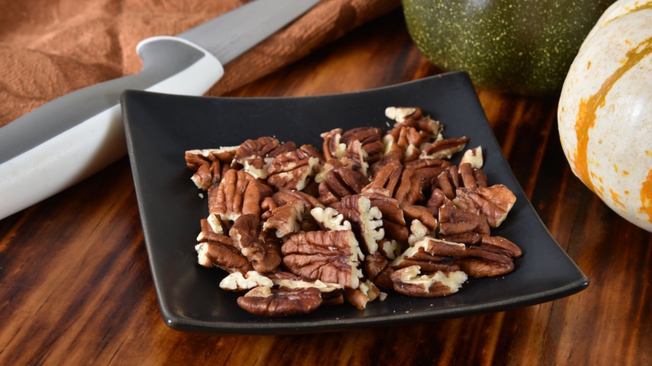 Chopped pecans in a small dish on a wooden cutting board.