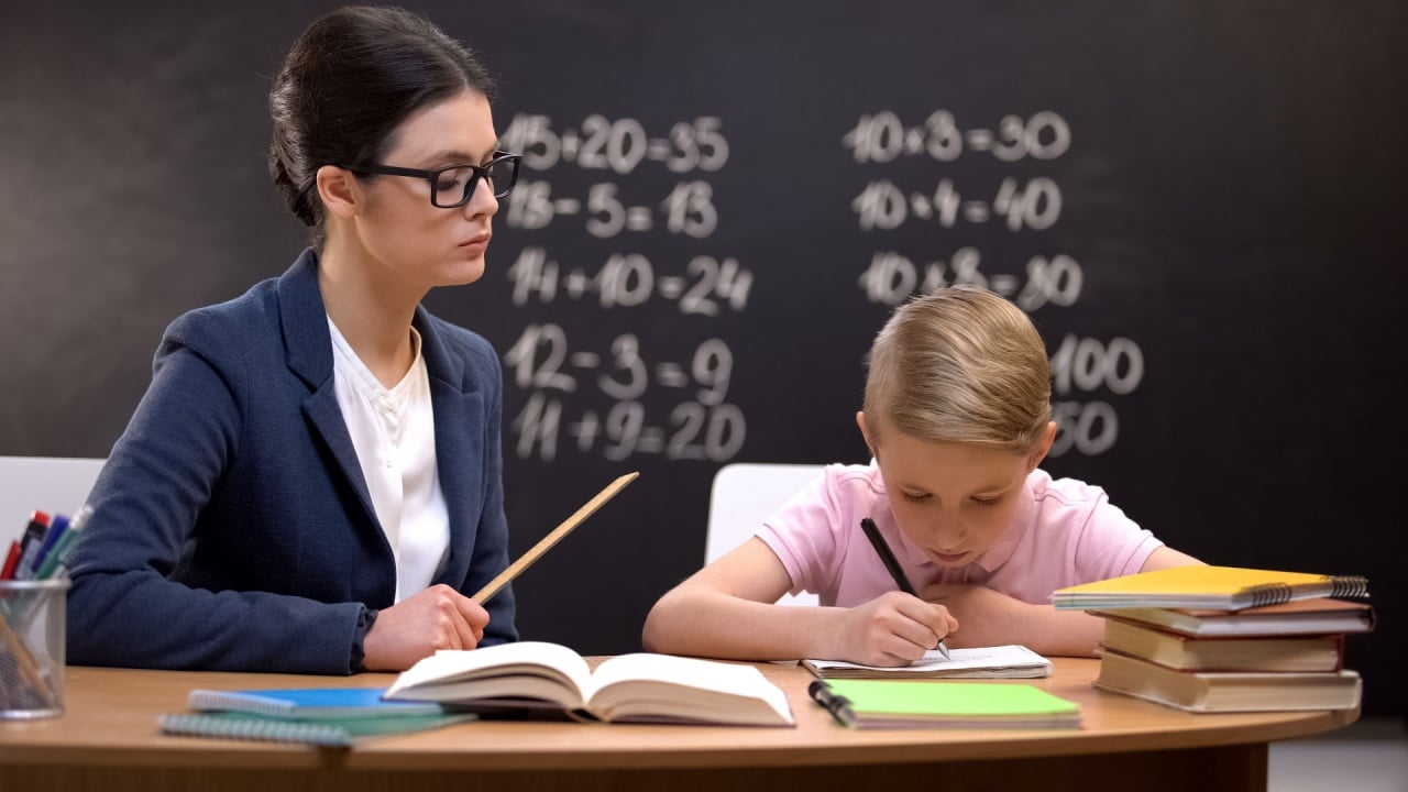 Strict teacher with ruler watching schoolboy doing his homework, control