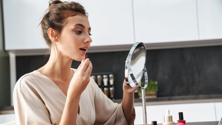Image of caucasian young beautiful woman in leisure clothes sitting by table at home and applying face makeup
