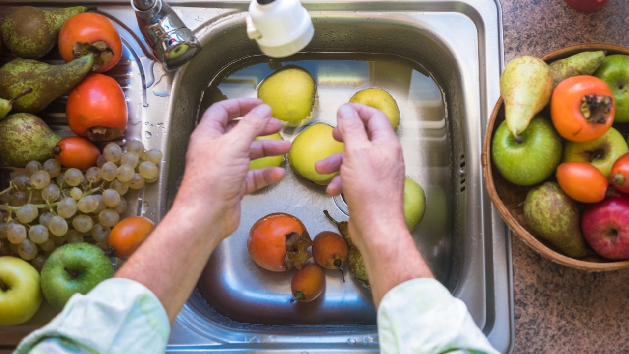 Top view of two human hands washing a mix of fresh fruit. Bright light from the window. A lot of fresh fruit around him