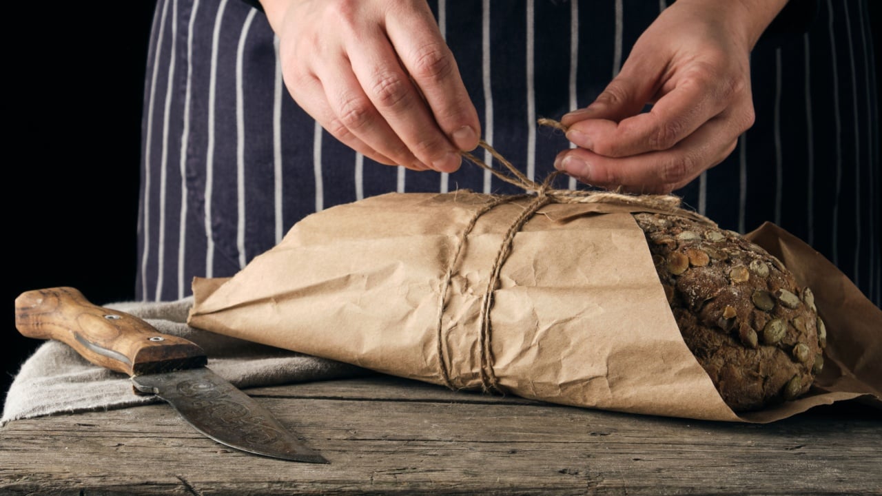 girl in black clothes wraps a whole baked loaf of bread in brown kraft paper and ties