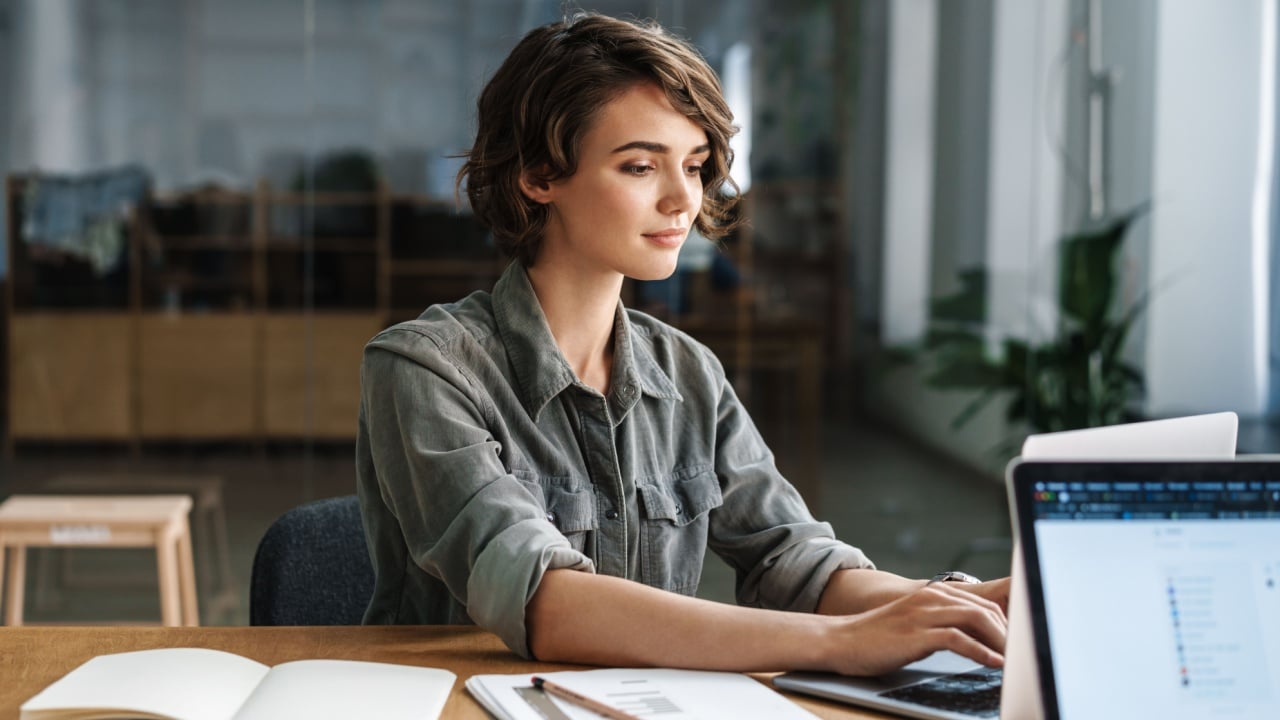 Image of young beautiful focused woman working with laptop while sitting at table in office