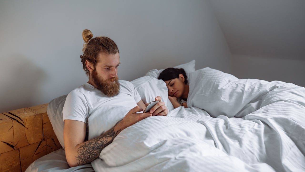 Man using phone in bed while his girlfriend sleeping next to him