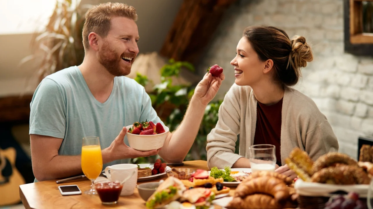 Happy couple having fun while eating breakfast at home. Man is feeding girlfriend with strawberries.