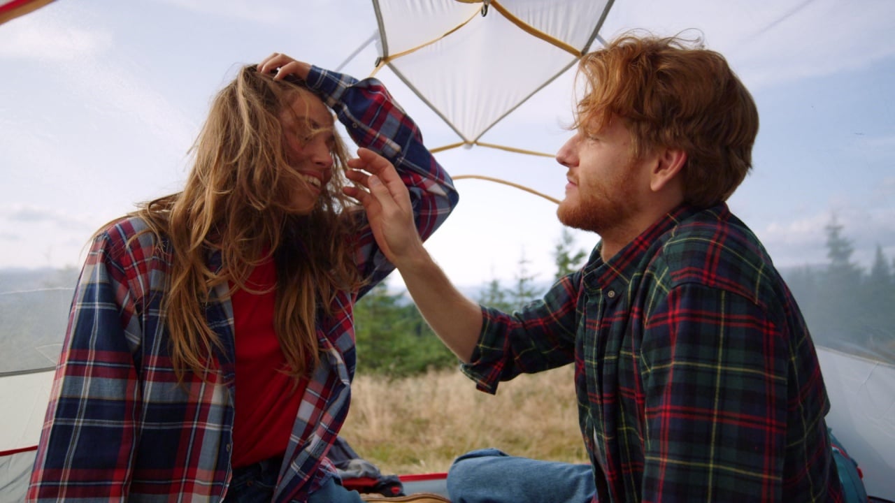 Affectionate man touching woman hair in tent after hike in mountains. Loving couple flirting with each other at summer weekend. Happy boyfriend and girlfriend cuddling together in camping tent