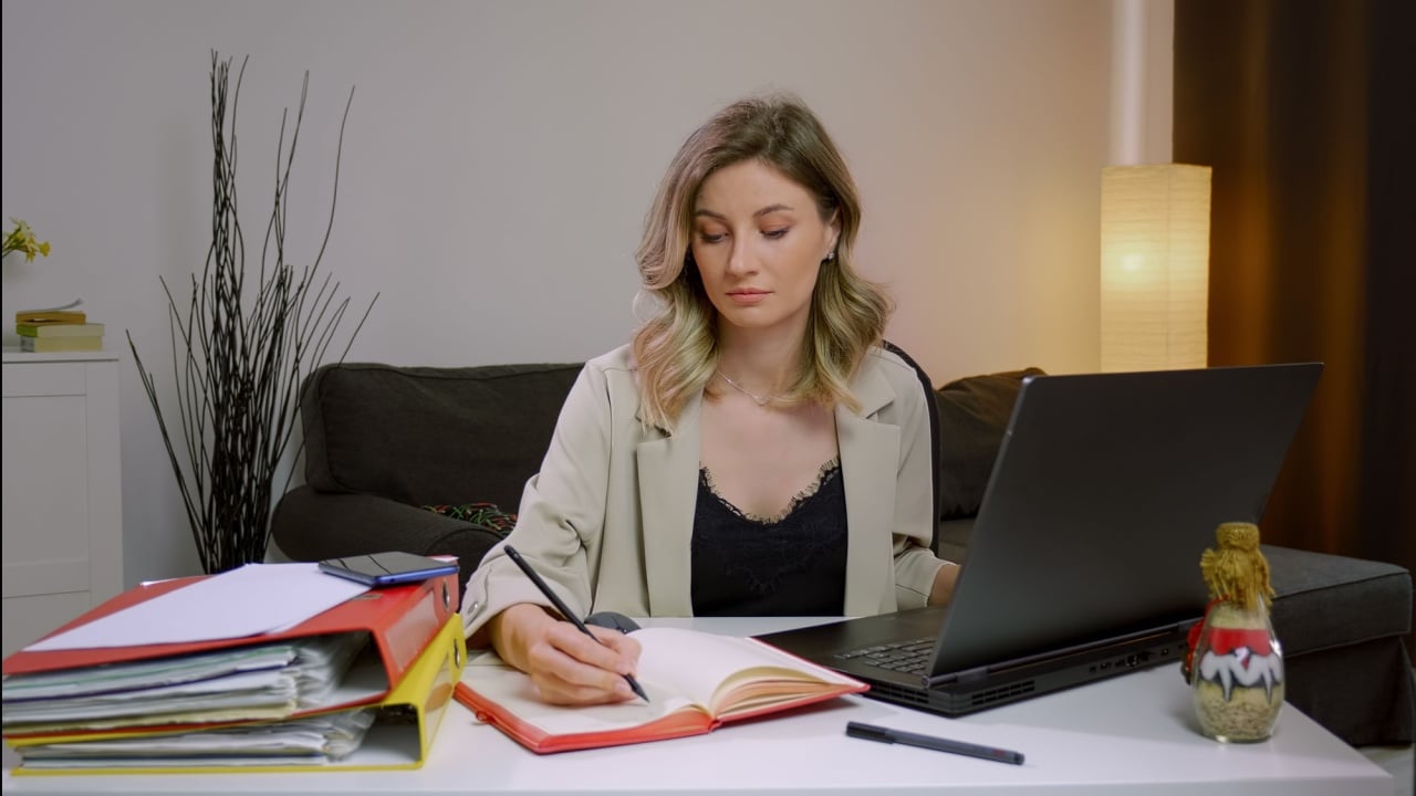 Portrait pretty focused business woman working on laptop in home office. Makes notes in notebook. Successful Caucasian woman concept. Office folders on table, modern house.