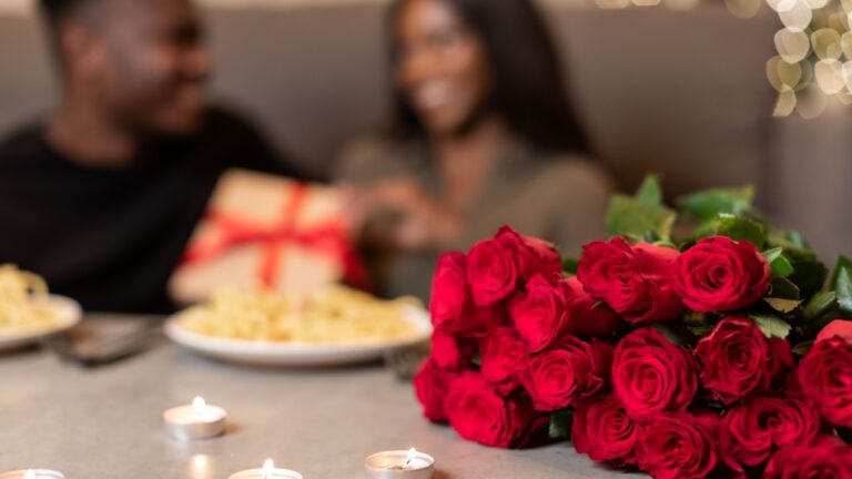 Valentine's Day Concept. African American Couple Celebrating Valentine Holiday Holding Gift Box Sitting At Table With Food And Candles Indoor. Selective Focus On Bouquet Of Red Roses. Shallow Depth
