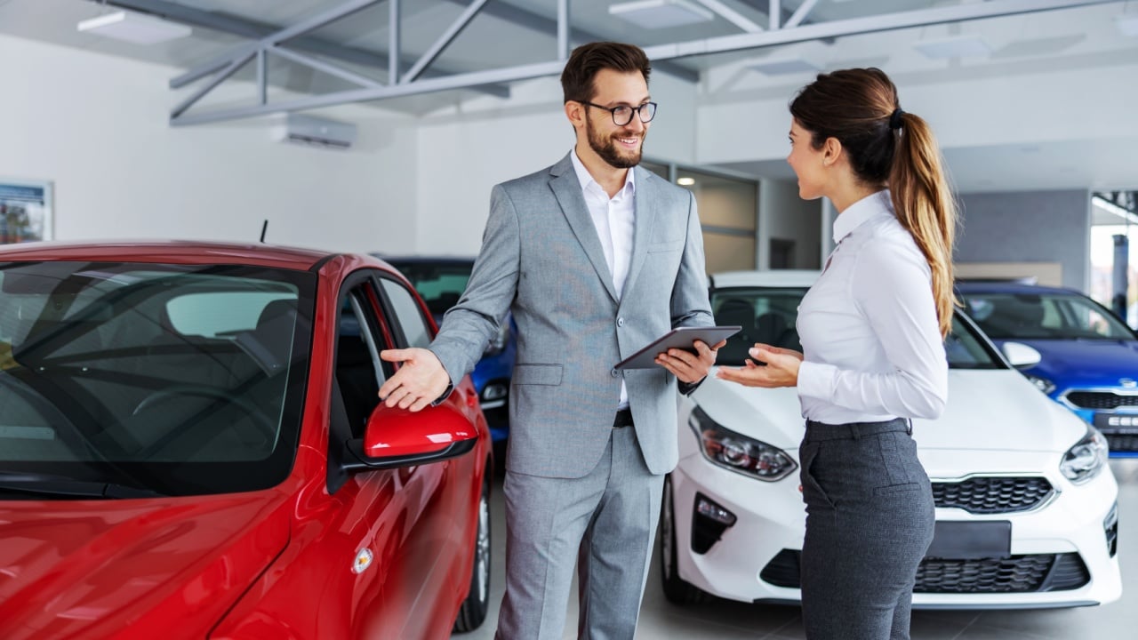 Smiling car seller standing in car salon with customer and showing around cars on sale.