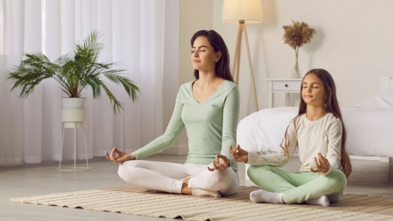 Family yoga. Beautiful young woman and her charming little daughter are smiling while doing yoga together at home. Family sits in lotus position on floor in living room. Mom teaches child to meditate.