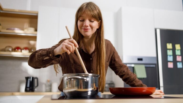 Cheerful young woman cooking and stirring food in saucepan at the kitchen