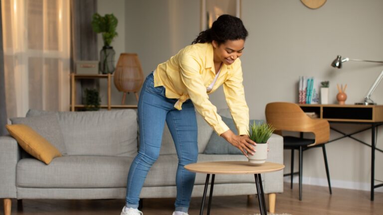 Smiling Black Female Putting Green Plant In A Pot On Little Table Decorating House And Cleaning Cozy Living Room At Home. Modern Interior Decoration And Housework Concept. Full Length