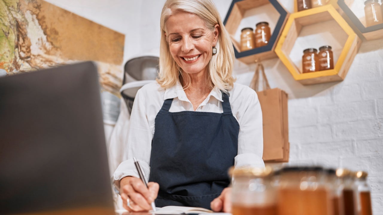 Laptop, small business and senior woman writing notes while doing inventory or online marketing. Happy, smile and elderly entrepreneur in a honey store working on admin or shop sales with a computer.