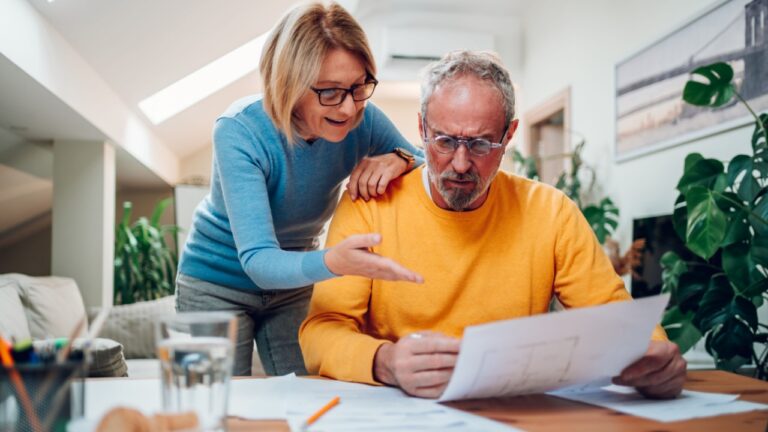 Senior married couple is actively discussing repairs in a new apartment or house while measuring the dimensions in the blueprints and sitting at the table and making notes.