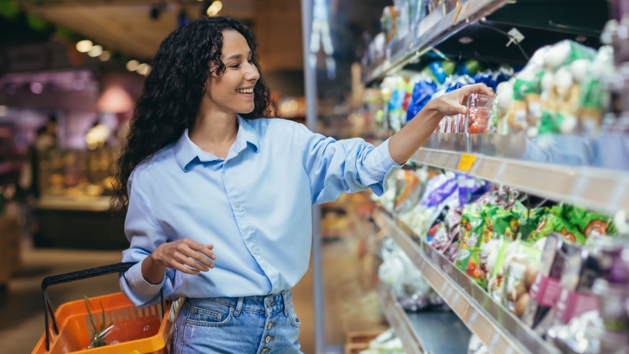 A young beautiful Hispanic woman buys cherry tomatoes in a supermarket. She stands smiling near the refrigerator, the vegetable department with a basket for groceries.