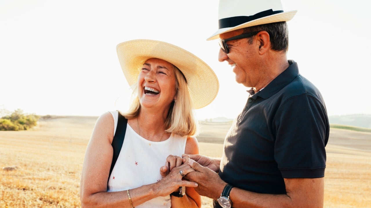 Senior cheerful couple holding hands, wear stylish clothes, sunglasses and hat, enjoying summer vacations together at field during sunset at Tuscany, Italy. Lifestyle, human relationships.