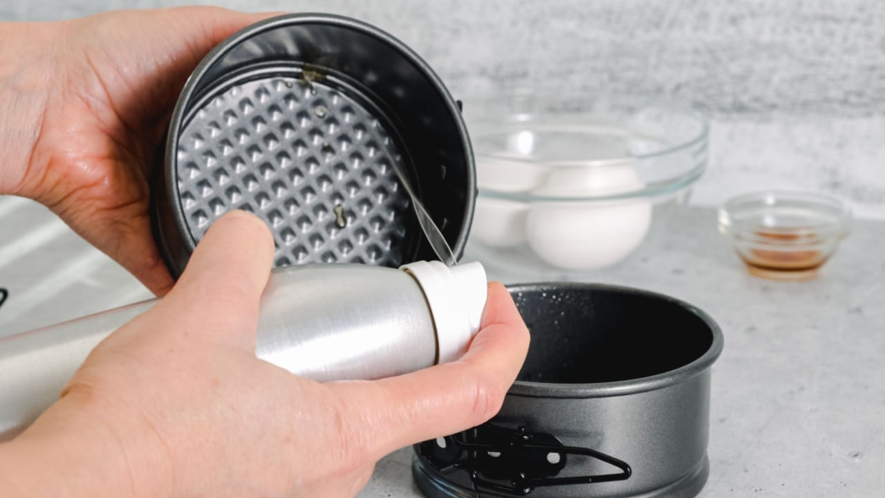 Woman using olive oil spray on a baking pan. Homemade mini cheesecakes preparation process, close-up ingredients