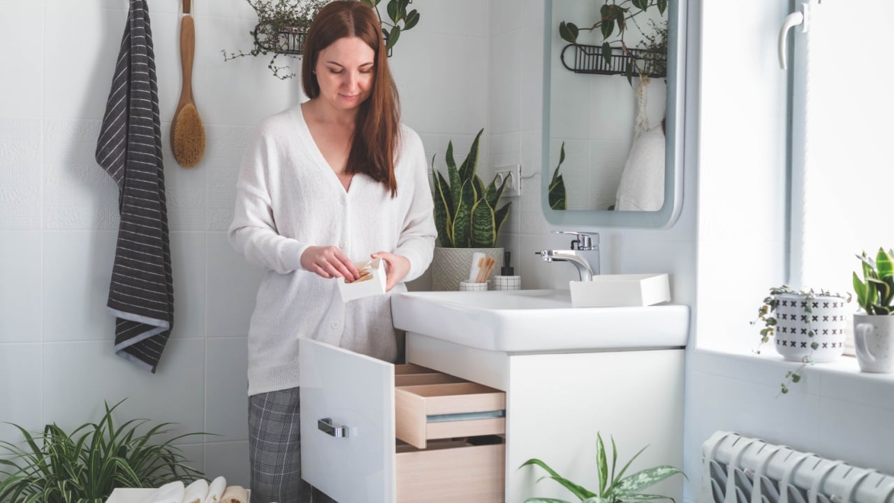 Happy young woman standing in front of open drawers of dressing table and taking out toiletries from drawer. Concept of saving space and organizing vanity table. Houseplants.